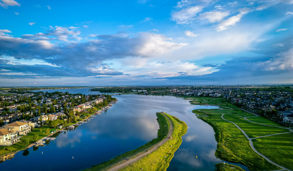 The image is an aerial photo of the City of Chestermere, showing Chestermere Lake with houses lining the shore, surrounded by green areas and roads. The sky is mostly blue with scattered clouds. 