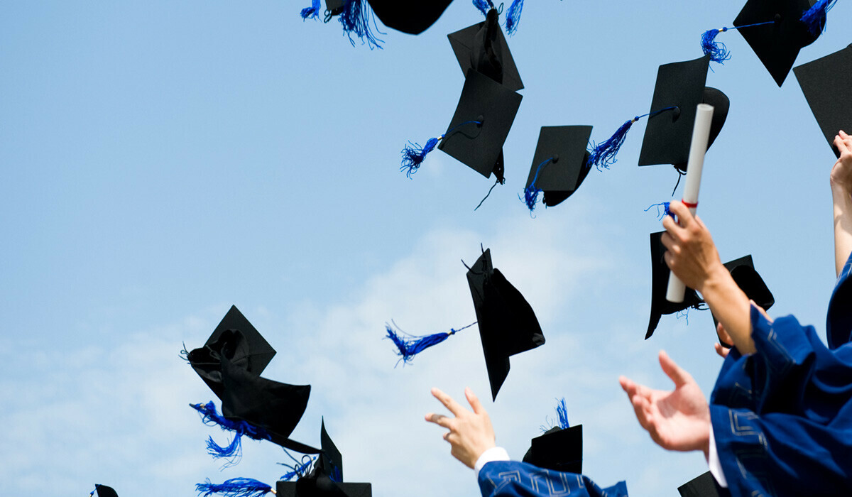 A group of graduates wearing blue gowns and throwing their black graduation caps with blue tassels into the air against a clear blue sky. Some hands are visible reaching up and one person is holding a rolled-up diploma.