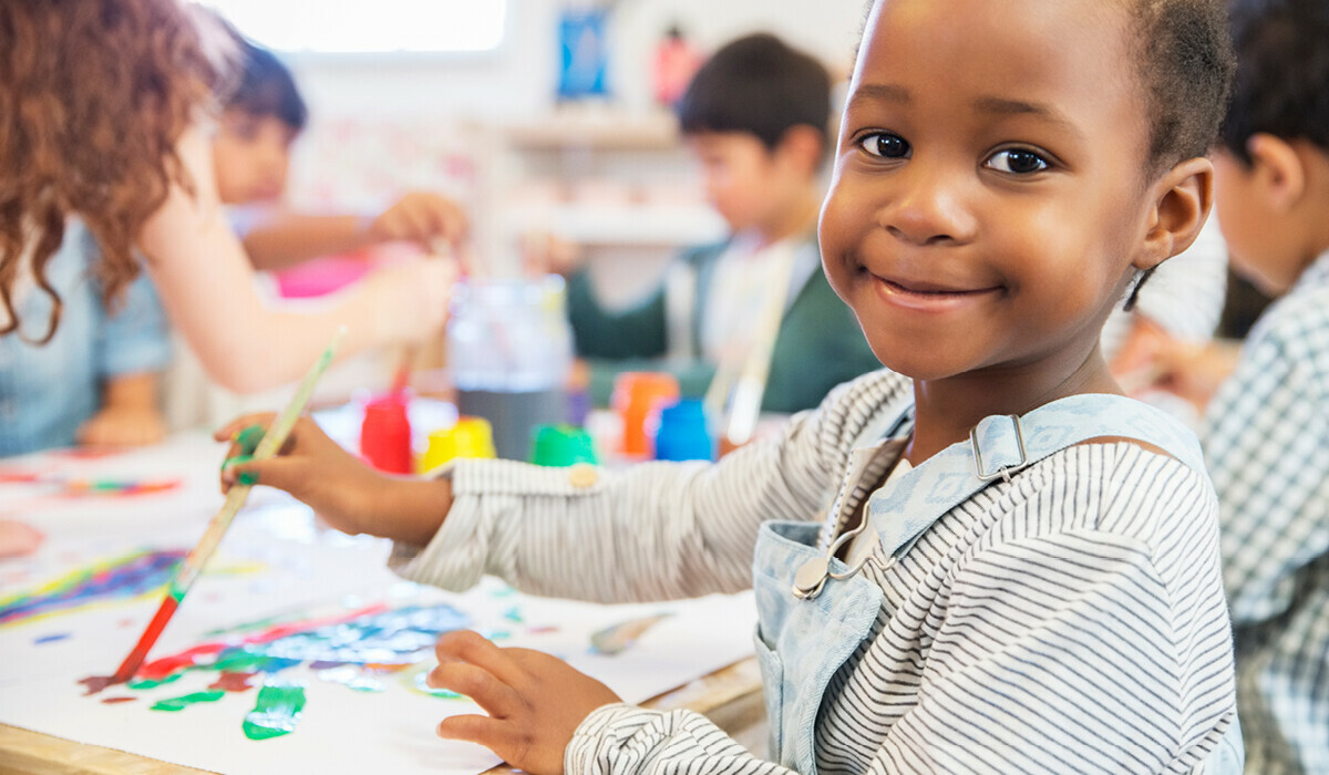 A smiling child in a striped shirt and overalls paints with a brush at a table surrounded by colourful art supplies and other children in a classroom.