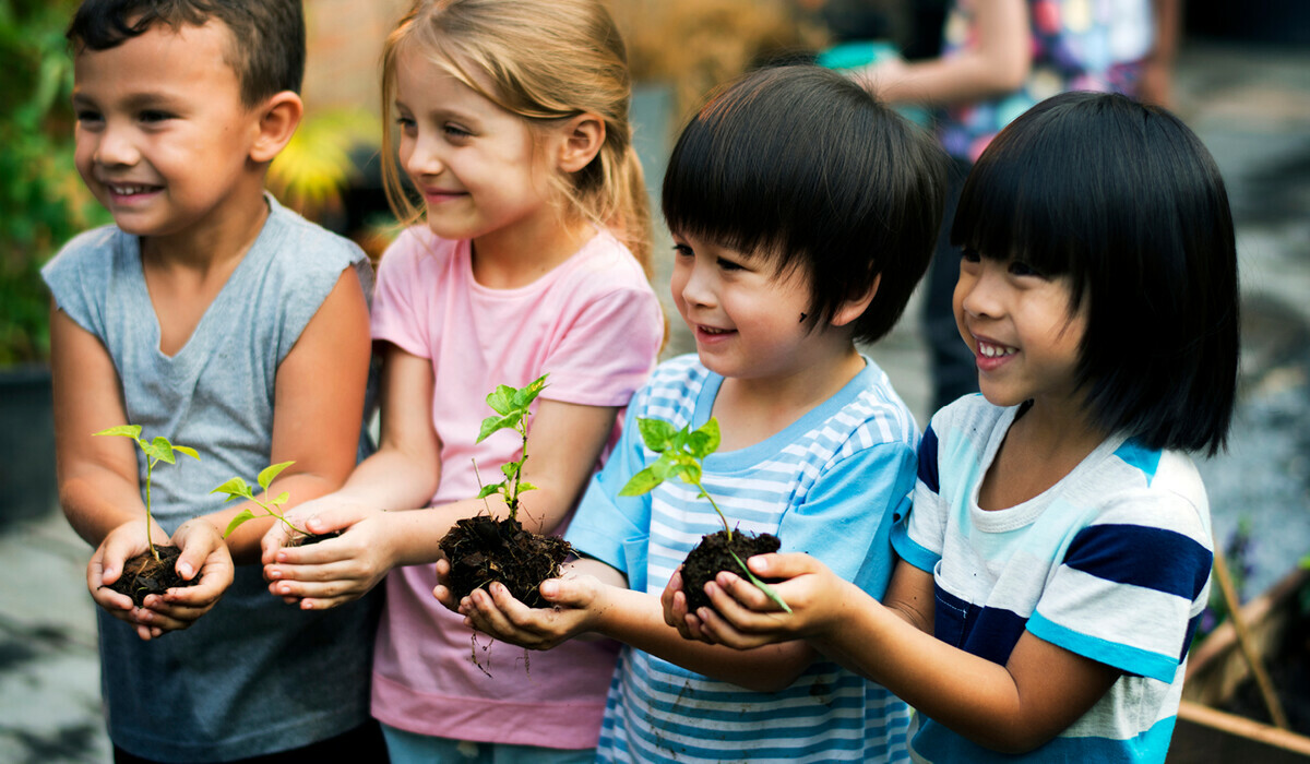 Three young children are holding small plants with soil in their hands, smiling.