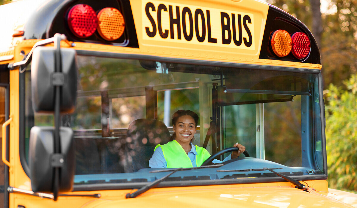 A smiling school bus driver wearing a safety vest sits behind the wheel of a yellow school bus