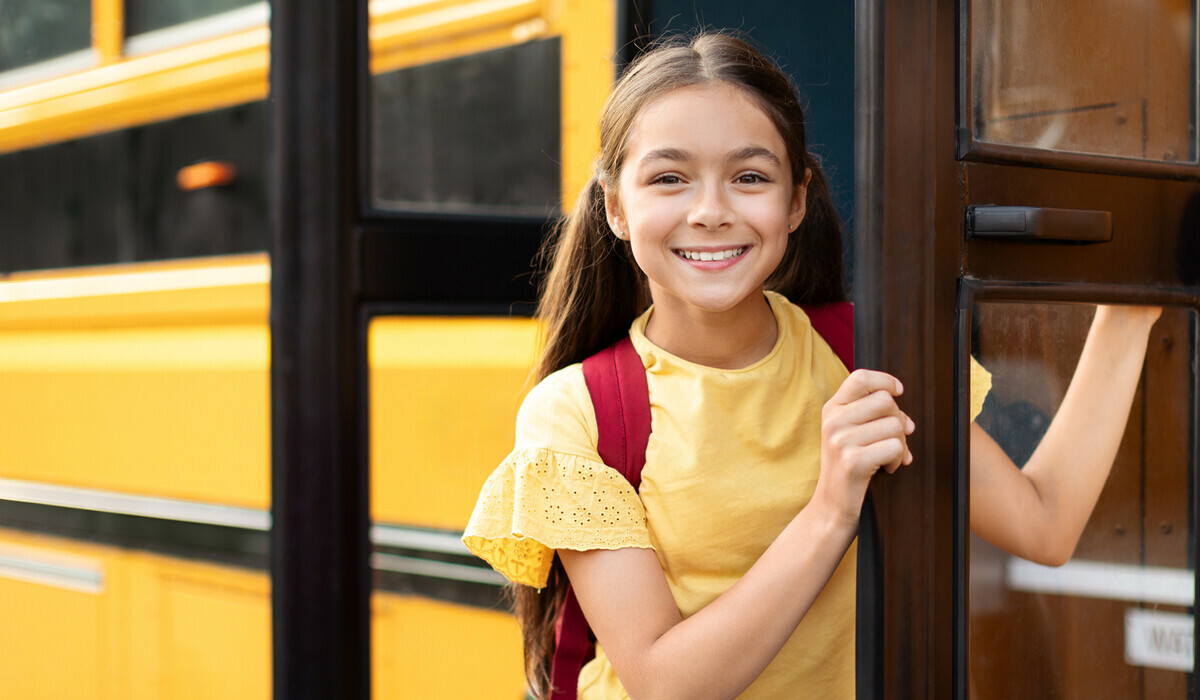 Young girl with brown hair, yellow shirt and red backpack, turning to smile as she boards the school bus.